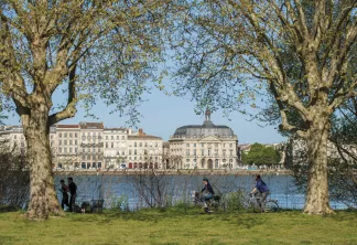 Vue panoramique de Bordeaux avec des arbres majestueux au premier plan, des cyclistes et promeneurs profitant du parc fluvial, et l'architecture historique se reflétant dans la Garonne.
