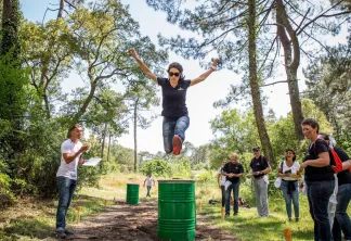 Une femme saute par-dessus un tonneau vert lors d'une épreuve d'entraînement en plein air dans une clairière forestière près de Bordeaux, entourée de spectateurs et de participants.