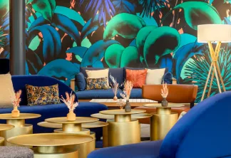Salon de luxe moderne avec canapés bleus courbes, tables basses cylindriques dorées, coussins décoratifs et un mur de fond orné d'une fresque tropicale aux feuillages verts et bleus