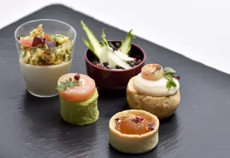 Amuse-bouches gastronomiques présentés sur ardoise noire : verre de mousse crémeuse garnie d'herbes, cylindre à base de pistache surmonté de saumon fumé, tartelette avec crème blanche et noix de Saint-Jacques, petit feuilleté au caramel, et bol décoratif 