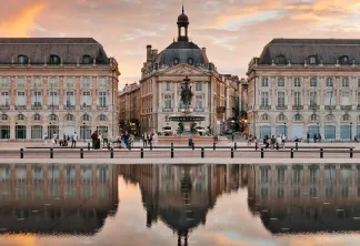 Place de la Bourse à Bordeaux avec ses bâtiments classiques du XVIIIe siècle se reflétant dans le miroir d'eau, ciel de coucher de soleil en arrière-plan et touristes sur la place