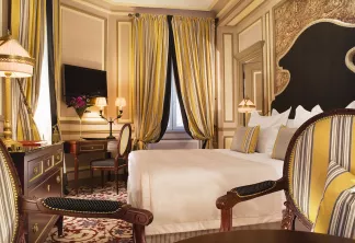 Chambre d'hôtel luxueuse à Bordeaux avec lit king-size en tissu rayé, palette de couleurs or et crème, mobilier en bois ouvragé, rideaux élégants et décoration française classique