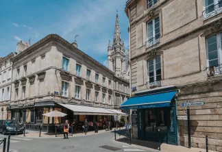 Vue de rue du centre-ville de Bordeaux montrant des bâtiments haussmanniens historiques, des commerces avec auvents, un clocher d'église en arrière-plan et le panneau de la station de métro Cité Mondiale par beau temps.