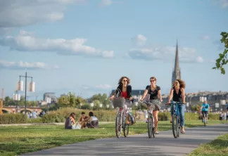 Trois femmes à vélo sur un chemin pavé dans un parc avec la silhouette de Bordeaux et sa cathédrale en arrière-plan, d'autres personnes profitant de l'espace vert.