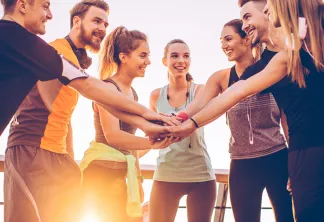 Un groupe diversifié de sept jeunes adultes vêtus de tenues de sport mettant leurs mains ensemble en cercle, souriant et célébrant à l'extérieur avec une lumière dorée en arrière-plan.