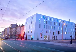 Bâtiment blanc moderne aux fenêtres illuminées au crépuscule à l'angle d'une rue de Bordeaux, à côté de bâtiments historiques aux teintes roses, avec des rails de tramway au premier plan