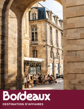 Voûte en pierre encadrant un immeuble de style haussmannien avec toit mansardé à Bordeaux, terrasse de café et piétons en bas