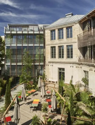 Cour contemporaine avec terrasse en bois, chaises longues, parasols et plantes en pot, bordée par un bâtiment moderne en verre à gauche et un immeuble classique en pierre à droite sous un ciel nuageux à Bordeaux.