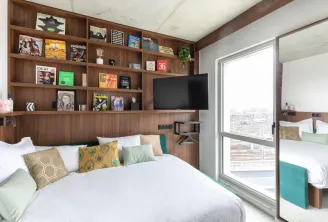Chambre d'hôtel moderne avec un lit queen-size, une unité de rangement murale en bois présentant des magazines et des objets de décoration, un téléviseur écran plat, des coussins d'accent colorés en or et turquoise, une grande baie vitrée donnant accès à 