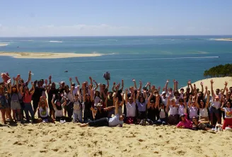Grand groupe de personnes célébrant sur une plage de sable avec les bras levés, dominant l'eau turquoise et les bancs de sable sous un ciel bleu clair