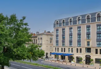 L'Hôtel Burdigala à Bordeaux, bâtiment de couleur beige et crème avec toit mansardé et fenêtres cintrées en mansarde, store bleu au rez-de-chaussée, entouré d'architecture ancienne et de verdure.