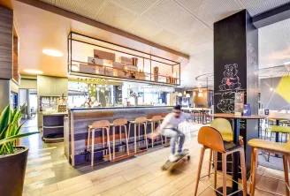 Intérieur contemporain du restaurant et bar de l'hôtel Novotel Bordeaux Lac avec comptoir et tabourets en bois, cuisine ouverte, éclairage suspendu, et une personne en skateboard traversant l'espace