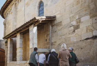 Un groupe de visiteurs rassemblés à l'entrée d'une ancienne église en pierre de style roman, dotée d'une porte en bois, d'une fenêtre en arc et d'une façade patinée sous un ciel bleu dégagé.
