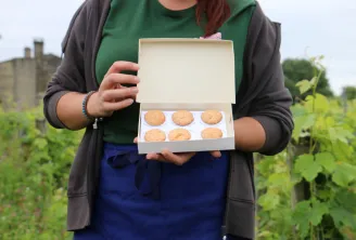 Femme tenant une boîte blanche ouverte contenant six biscuits faits maison dans un jardin, présentant les produits culinaires locaux de la région de Saint-Emilionnais.