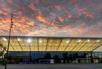 Centre de congrès moderne en verre et acier avec toit illuminé au crépuscule, surplombé d'un ciel dramatique aux teintes orangées et roses, situé à Bordeaux