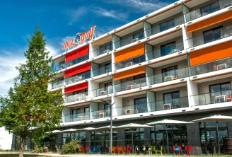 Immeuble d'appart-hôtel moderne aux balcons colorés orange et rouge, arborant le logo Mer & Golf sur la façade, situé à Bruges près de Bordeaux avec terrasse au rez-de-chaussée et mobilier extérieur coloré