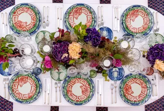 Table ovale élégamment dressée avec six couverts ornés d'assiettes aux motifs rouges et verts, verres transparents, serviettes blanches et couverts argentés, agrémentée d'une composition florale luxuriante aux fleurs pourpres, roses et crème.