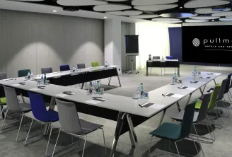 Salle de conférence moderne du Pullman Bordeaux Lac avec une table en U, des chaises colorées, des bouteilles d'eau, des blocs-notes, des luminaires au plafond et un grand écran affichant le logo Pullman Hotels.