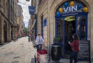 Une femme à vélo avec une remorque cargo passe devant une cave à vin 'Vin Cousin & Compagnie' à devanture bleue ornée de casques colorés, située sur une belle rue pavée de Bordeaux.