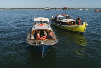 Deux bateaux de pêche avec des passagers amarrés dans des eaux calmes, avec un paysage côtier et des bâtiments visibles en arrière-plan lors d'un rallye nautique.