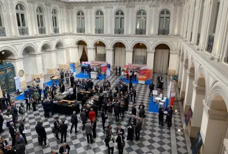 Vue aérienne d'une grande conférence ou événement de réseautage dans une cour classique élégante au sol en damier, réunissant des participants en tenue professionnelle, des stands d'exposition bleus et une architecture blanche ornée avec des fenêtres cint