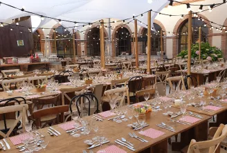 Un lieu de restauration élégant en plein air à Bordeaux avec de longues tables en bois dressées pour un événement formel, présentant des sets de table à carreaux roses, des verres en cristal et des couverts, sous une toile rétractable avec guirlandes lumi