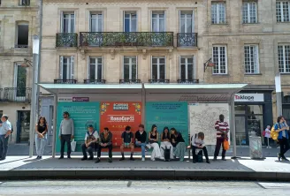 Personnes attendant à un abri-bus contemporain devant un immeuble haussmannien classique à Bordeaux, avec des affiches informatives incluant l'événement RoboCup 2023 et une carte de la ville.