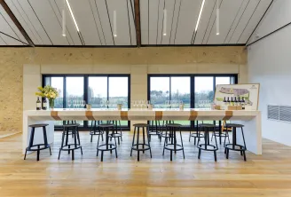 Salle de dégustation de vin moderne du Château La Garde à Bordeaux avec un long comptoir blanc, des chaises hautes noires, de grandes baies vitrées donnant sur les vignobles, des murs en liège et un plafond contemporain aux lignes épurées.