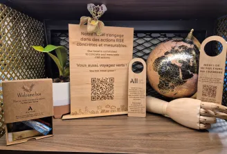 Vitrine de chambre d'hôtel présentant des initiatives de tourisme durable avec panneaux en bois sur les engagements RSE, globe en liège, boîte de bienvenue, plante décorative et codes QR pour les informations de voyage écologique.