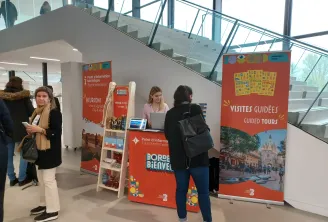 Point d'information touristique du centre d'accueil de Bordeaux avec des visiteurs consultant le personnel à des kiosques rouges présentant les visites guidées et informations sur la ville, intérieur moderne avec escaliers et garde-corps en verre visibles