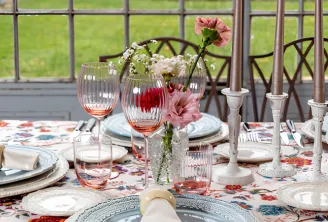 Table de repas élégante dressée avec nappe à motifs floraux, couverts raffinés aux assiettes décoratives, compositions florales avec œillets roses et gypsophile, chandeliers et verres rosés, vue sur le jardin par grandes fenêtres.
