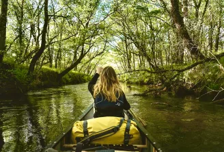 Femme en kayak naviguant dans un cours d'eau bordé d'arbres avec un sac étanche jaune fixé au bateau, pagayant dans des eaux calmes entourées de végétation luxuriante.