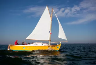 Voilier en bois jaune avec voiles blanches naviguant sur l'océan, numéro d'immatriculation AC D82516 visible sur la coque, plusieurs personnes à bord, ciel bleu nuageux en arrière-plan