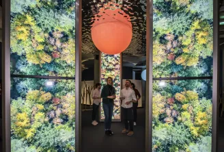 Trois visiteurs se tiennent dans un couloir de dégustation sensorielle moderne avec des panneaux verticaux illuminés affichant des vues aériennes de forêt de chaque côté et une grande sphère orange luminescente suspendue au plafond.
