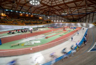 Vélodrome couvert du Stadium de Bordeaux Lac avec des cyclistes en compétition sur la piste inclinée, terrain d'athlétisme avec haies et équipements d'entraînement au centre, gradins de spectateurs visibles en arrière-plan.
