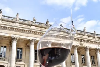 Un verre de vin rouge levé devant un bâtiment classique en pierre avec colonnes et statues, représentant la culture vinicole de Bordeaux.
