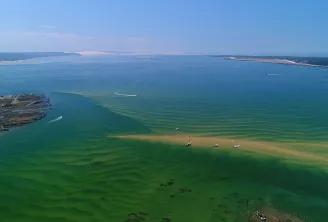 Vue aérienne du Bassin d'Arcachon montrant des eaux turquoises peu profondes, un grand navire de croisière amarré à gauche, des voiliers dispersés et des plages de sable entourées de forêts de pins.