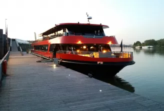 Un bateau de croisière fluviale rouge et noir amarré à un ponton en bois à Bordeaux, illuminé en soirée, avec la Garonne et l'architecture urbaine en arrière-plan.