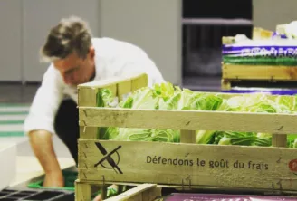 Un homme en tenue de chef blanc organisant de la laitue fraîche dans des caisses en bois portant l'inscription 'Défendons le goût du frais' dans une cuisine professionnelle.