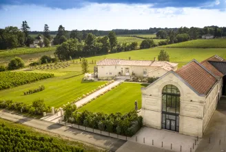 Vue aérienne du Château La Garde à Bordeaux, montrant un bâtiment moderne en pierre avec baies vitrées cintrées, jardins aménagés et vignobles étendus entourés de champs verts et d'arbres sous un ciel bleu.