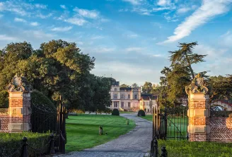 Portail d'entrée du Château Giscours à Bordeaux, avec piliers en brique ouvragée, portail en fer forgé, pelouse soignée et le château visible au loin sous un ciel bleu