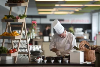 Chef en uniforme blanc et toque préparant un plat au comptoir d'une cuisine moderne, avec des produits frais exposés sur des étagères en bois et un panier en osier à proximité
