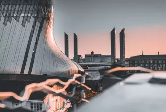 Bâtiment moderne aux lignes courbes et façade vitrée longeant l'architecture historique au bord de l'eau à Bordeaux, avec tours jumelles et bâtiments traditionnels au coucher de soleil