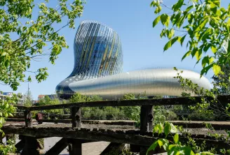 Bâtiment contemporain aux formes courbes en verre et acier avec architecture ondulante, encadré par la végétation et un ancien pont métallique industriel au premier plan