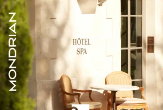 Entrée du spa de l'hôtel Mondrian Bordeaux, avec des chaises en bois élégantes et une petite table ronde dans une cour ensoleillée aux murs en pierre crème et lierre grimpant.
