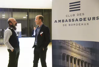 Deux hommes en tenue professionnelle discutant au Club des Ambassadeurs de Bordeaux, avec la signalétique du club visible sur le mur et une photographie d'un bâtiment classique affichée en dessous.