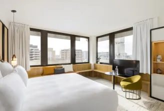 Chambre d'hôtel contemporaine avec lit double, coin fenêtre avec banquette jaune moutarde, télévision écran plat et baies vitrées donnant sur la skyline de Bordeaux