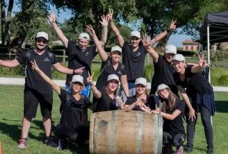 Une équipe de personnes vêtues de t-shirts noirs et de casquettes blanches posant ensemble en extérieur devant un tonneau en bois, les bras levés en signe de célébration dans un cadre rural.