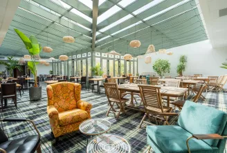 Salle de restauration contemporaine sous véranda vitrée avec mobilier en bois, plantes en pot, luminaires suspendus tressés et fauteuils colorés sur moquette à carreaux