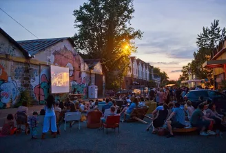 Projection de cinéma en plein air au coucher du soleil dans une cour publique entourée de bâtiments historiques en brique ornés de street art, où une foule diversifiée de personnes assises sur des chaises colorées regarde un film projeté sur un écran blan